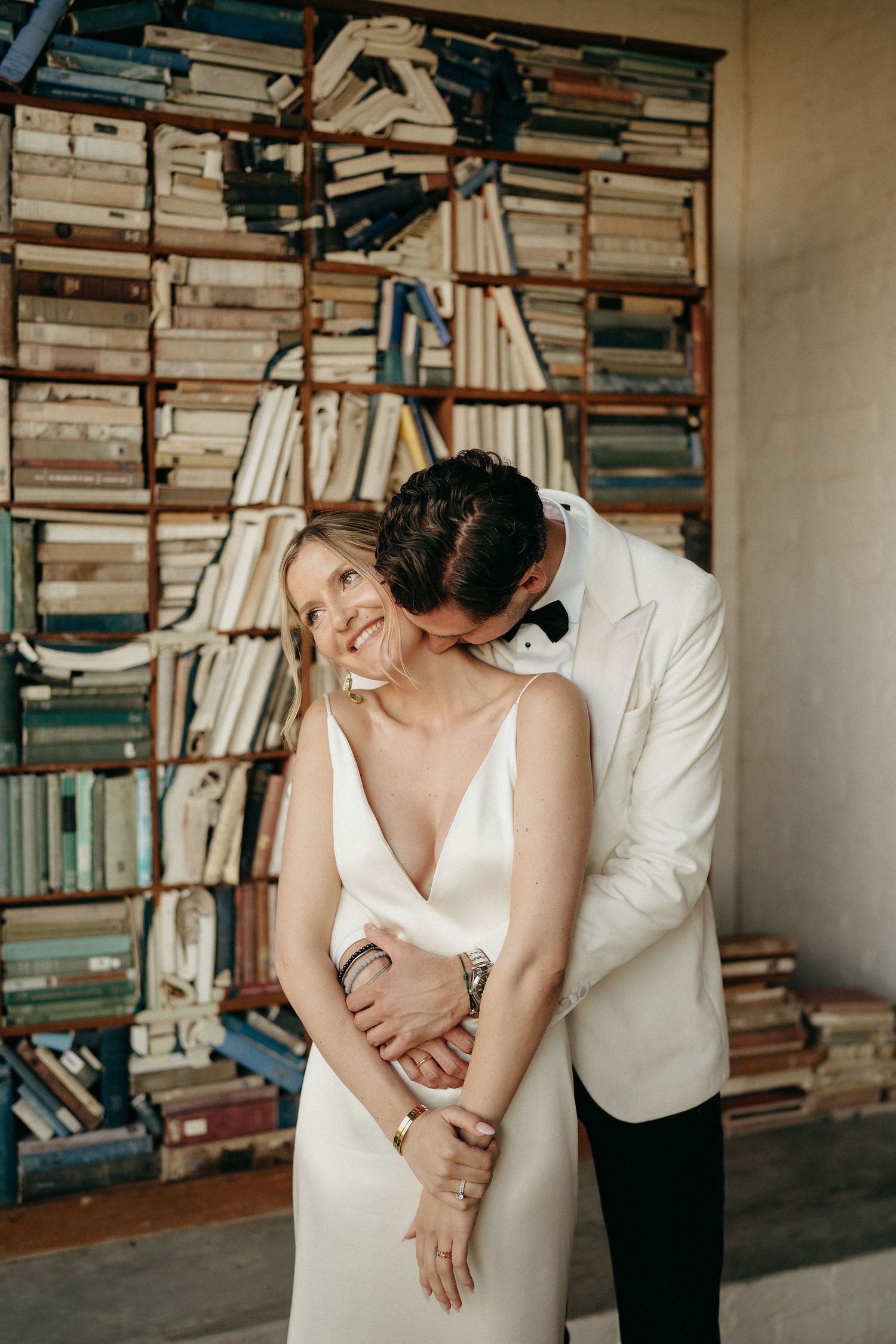 A romantic couple embraces and laughs together in front of a wall of bookshelves filled with vintage books.
