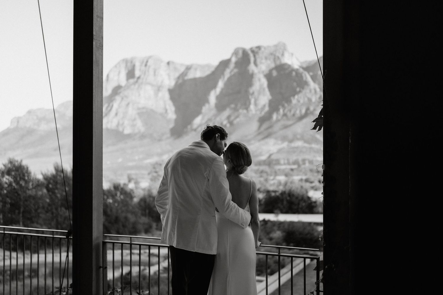 A couple embraces while overlooking a scenic mountain landscape from a balcony in black and white photography.