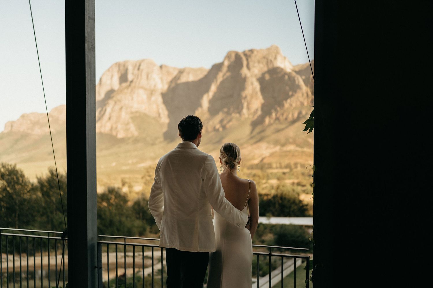 Two figures stand on a balcony overlooking scenic mountain views at sunset.