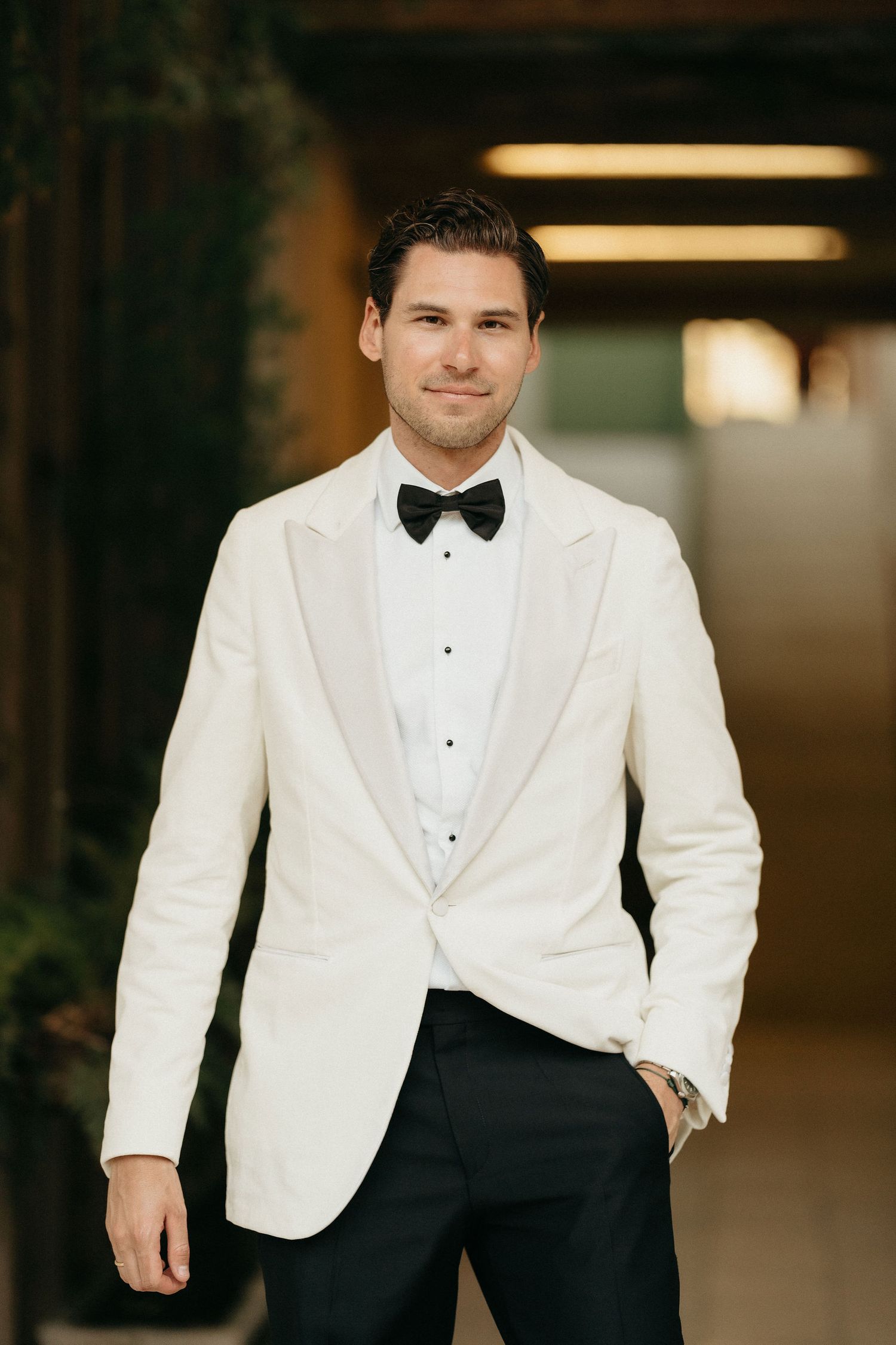 A groom in a classic white tuxedo jacket and black bow tie poses for formal wedding photos in a dimly lit venue.