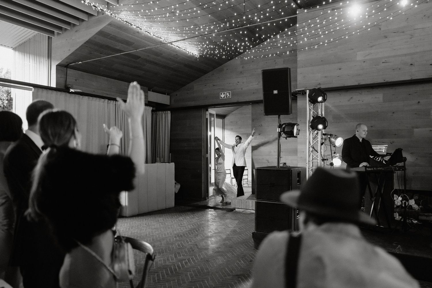 A black and white panoramic photo of people dancing at a wedding reception with string lights overhead.