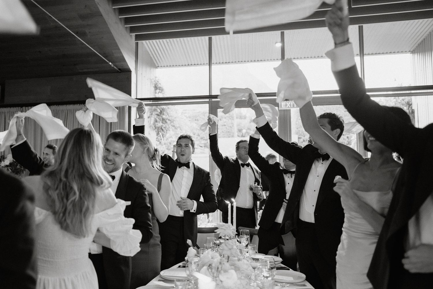 Wedding guests raise their napkins in celebration during a joyful reception dinner in black and white photography.
