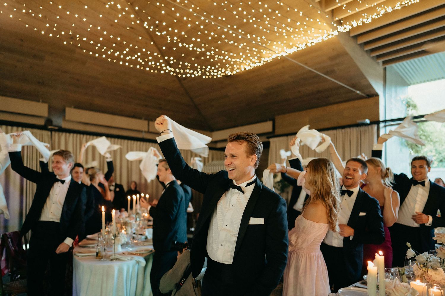 Wedding guests dance together under a ceiling adorned with twinkling string lights at an elegant reception venue.