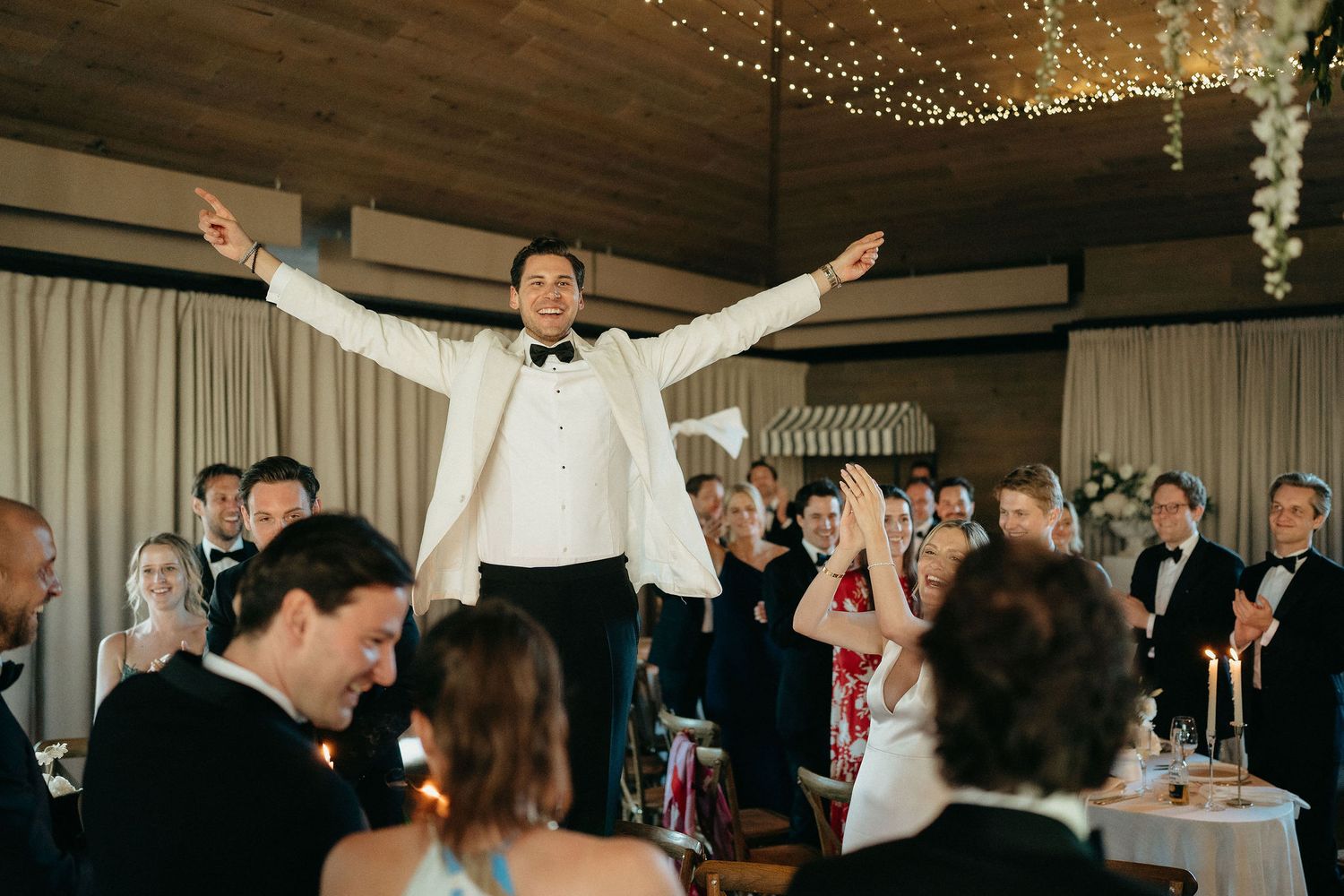 Wedding guests dance and celebrate on the dance floor during an indoor reception party with string lights overhead.