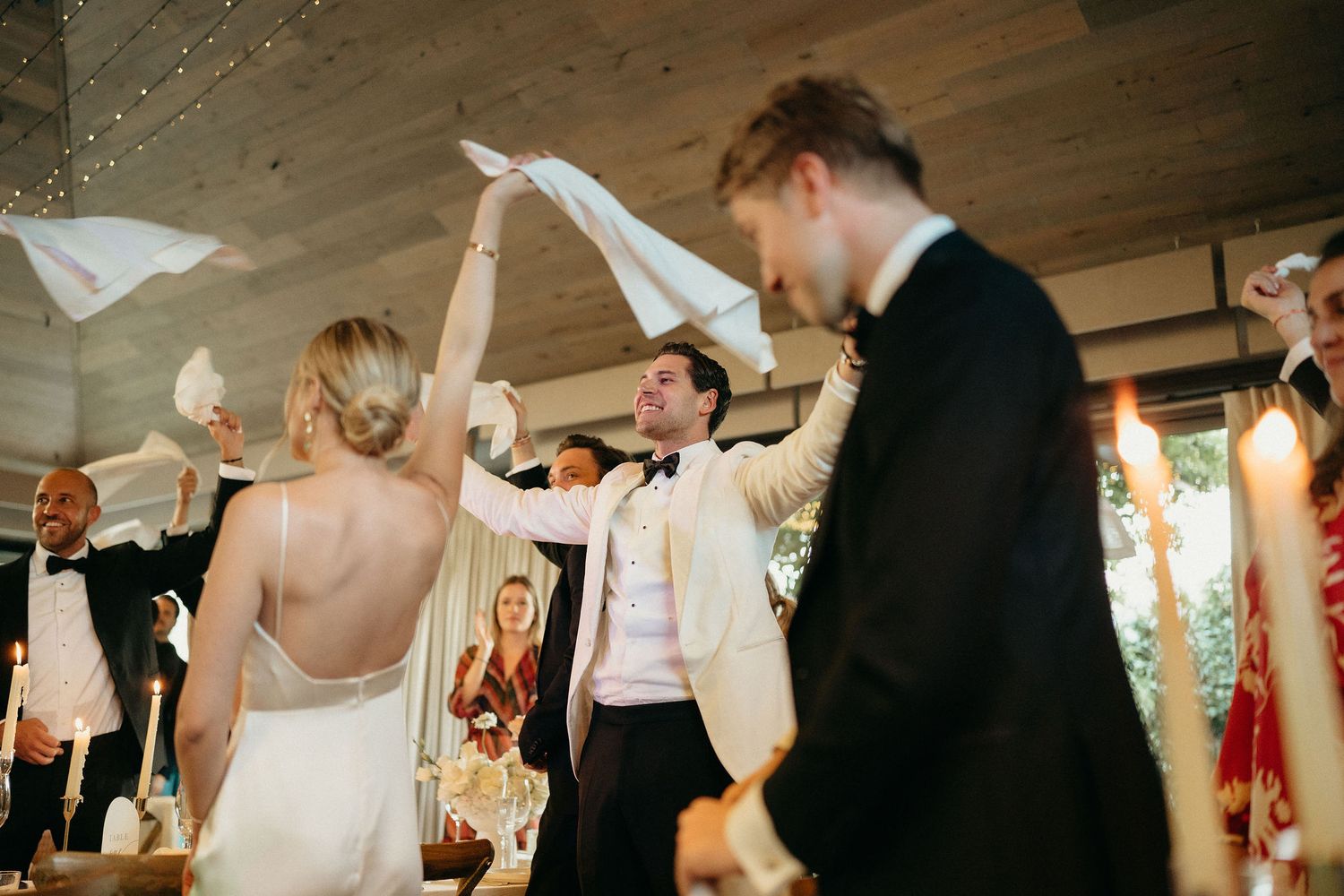 Wedding guests wave white napkins in celebration during a lively reception party in a rustic venue.
