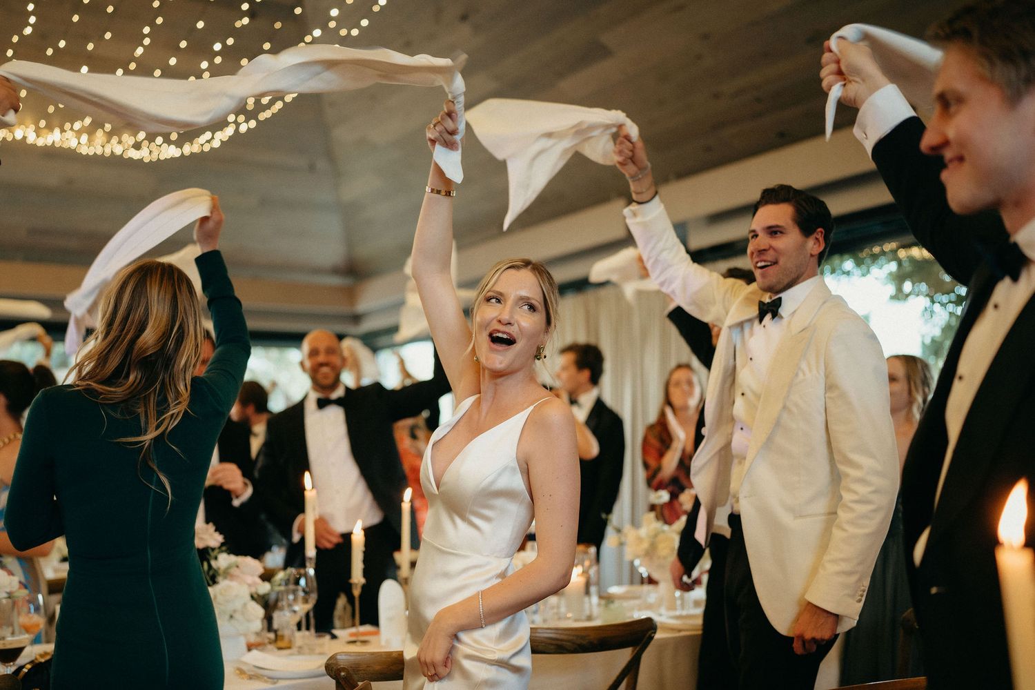 A group of wedding guests dancing and celebrating on a dance floor under twinkling string lights at a reception.