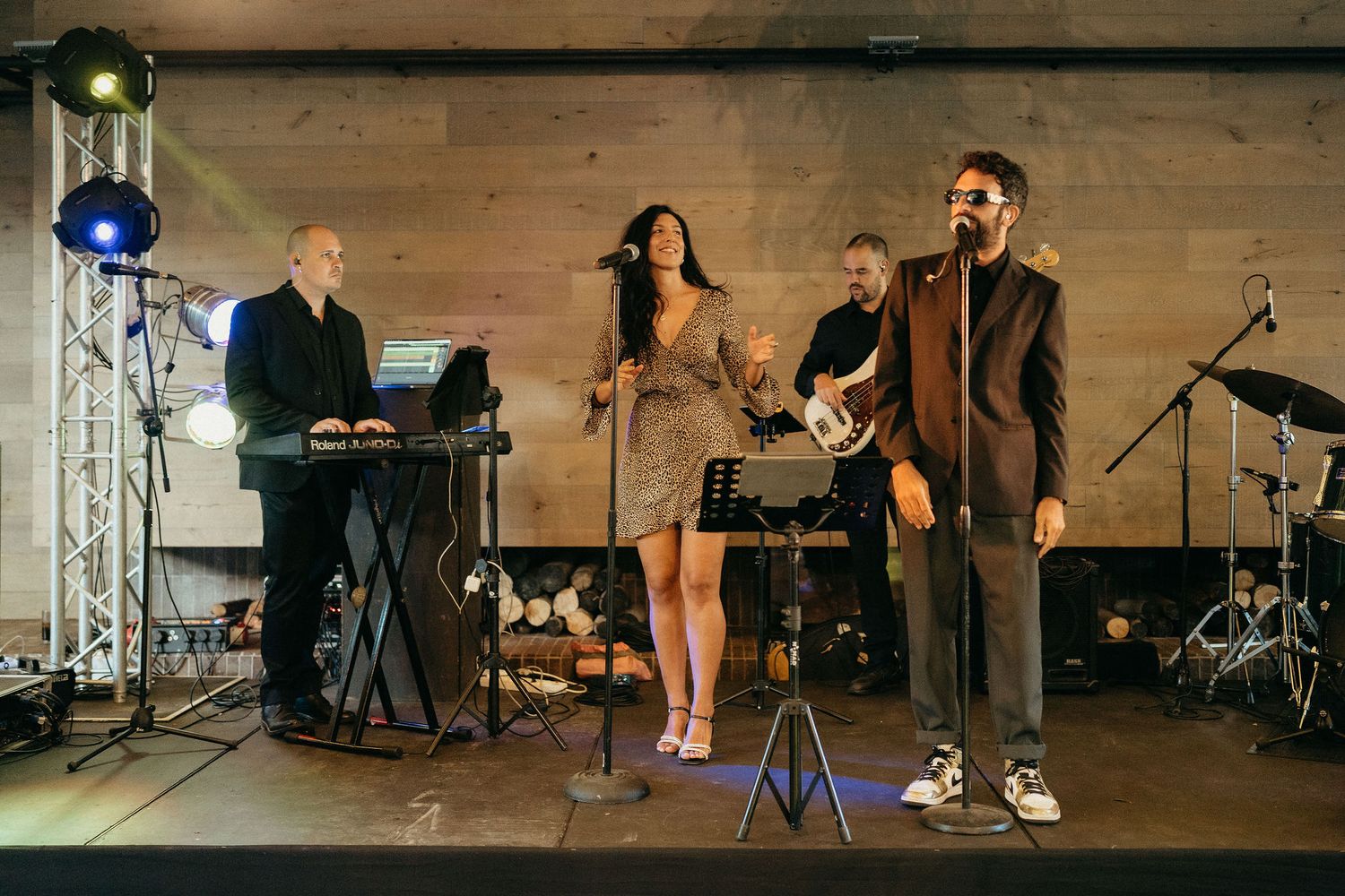 Musicians perform on stage with instruments and microphones in a rustic indoor venue with wooden beams and lighting.