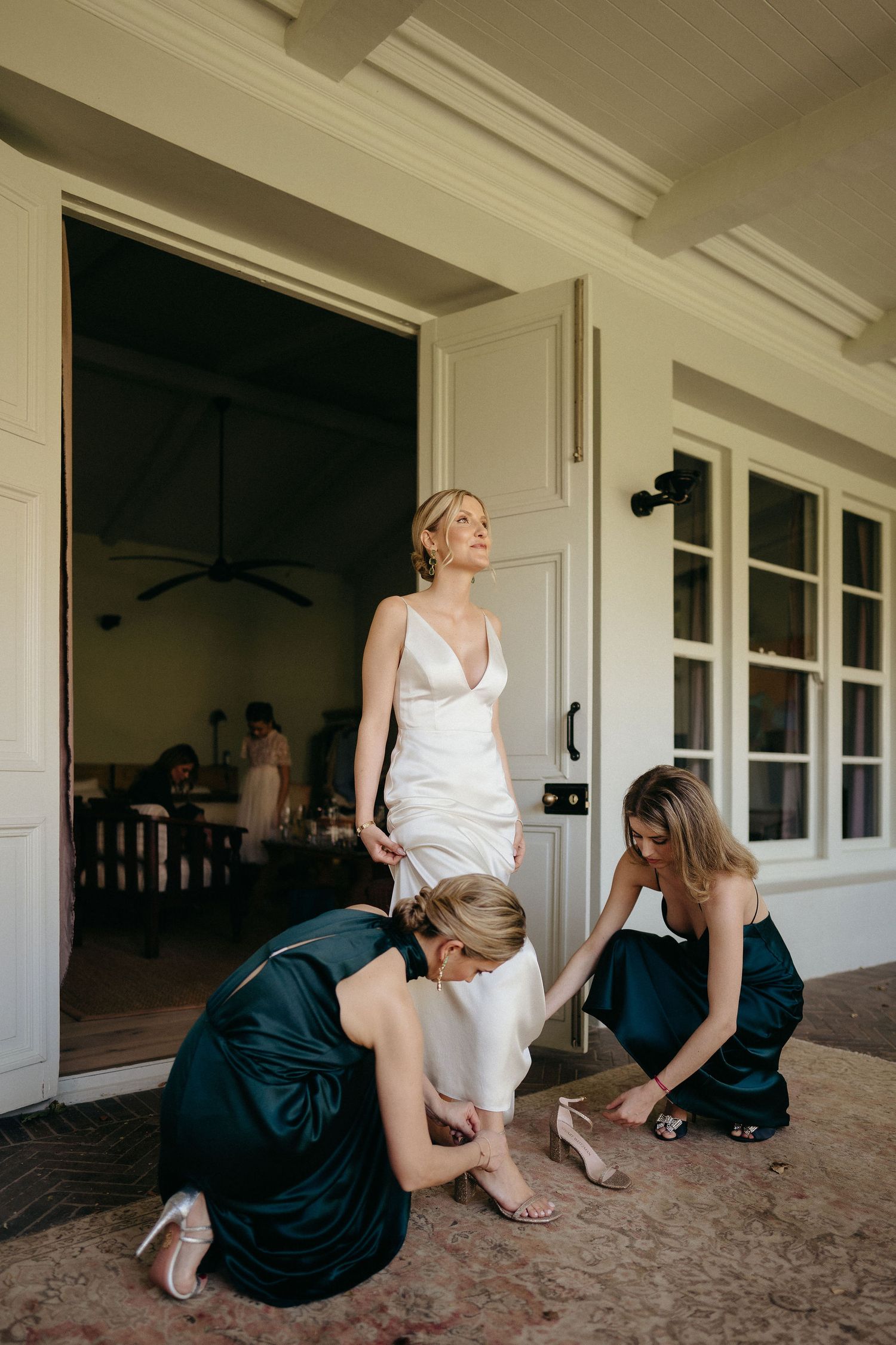 Wedding party members in teal dresses help adjust the bride's white gown on a covered porch.