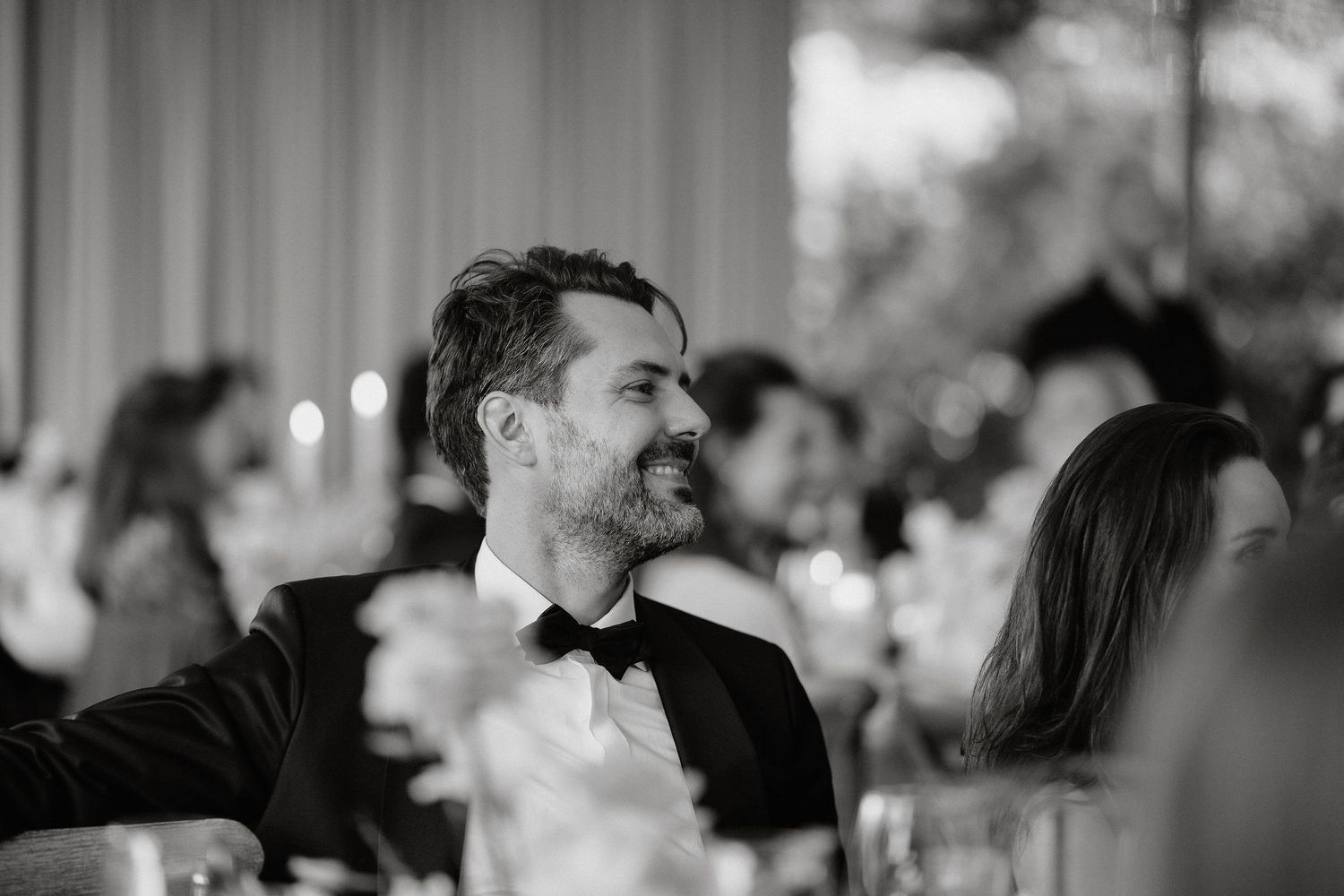 A guest in a black bowtie smiles while seated at an elegant wedding reception dinner table.