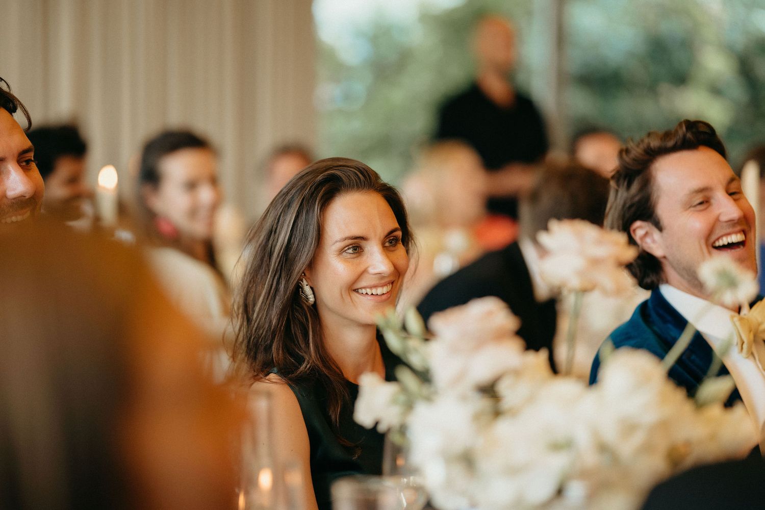 Wedding guests share joyful laughter during an elegant indoor reception with floral centerpieces.
