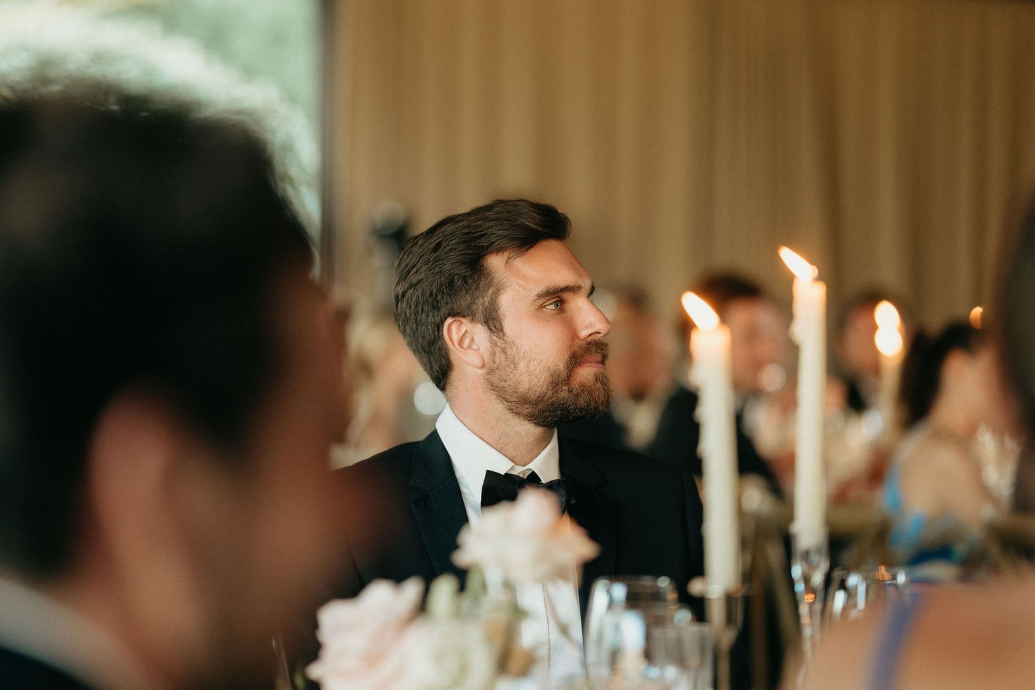 A guest in formal attire listens intently during a candlelit wedding reception dinner.