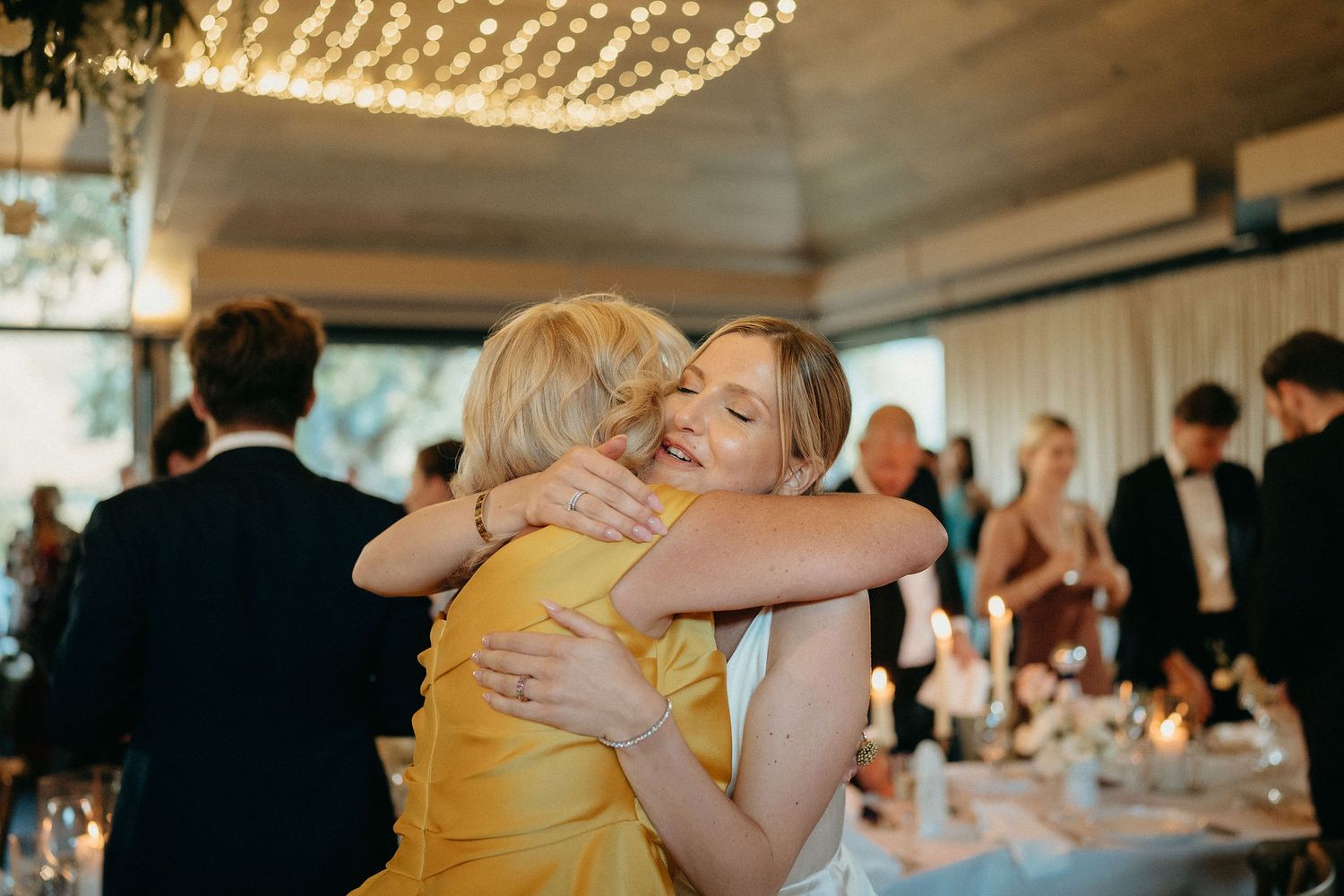 Two people share an emotional embrace during what appears to be a wedding reception with string lights overhead.