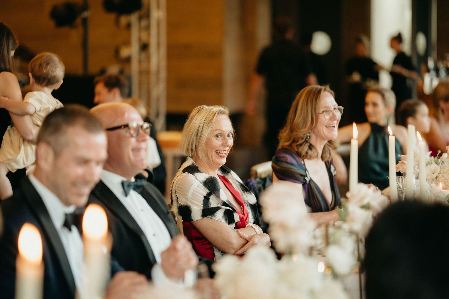 Wedding guests in formal attire laugh and smile during an elegant candlelit reception dinner.