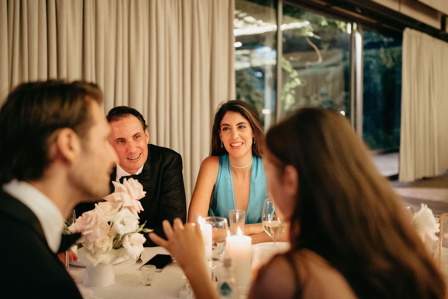 Wedding guests enjoy an elegant candlelit dinner reception with white floral centerpieces and formal table settings.