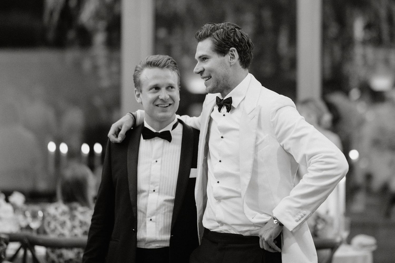 Two men in formal attire share a candid moment at an event, one wearing a black tuxedo and one in white.