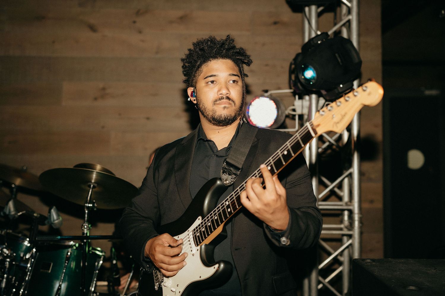 Musician plays electric guitar on stage with drum kit visible in background during live performance.