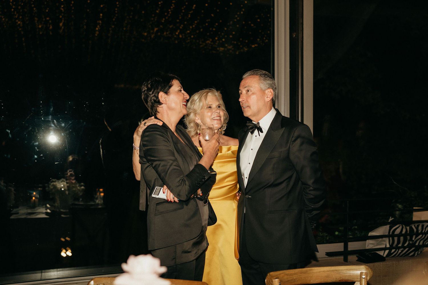 Three people in formal attire share a laugh together at an evening event.