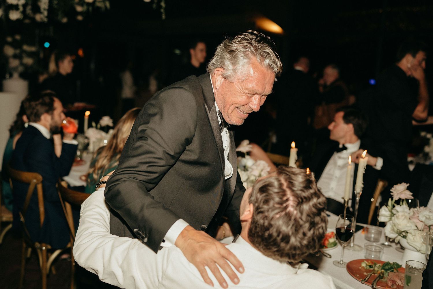 A person in a grey suit jacket leans over a dining table at an evening reception event.
