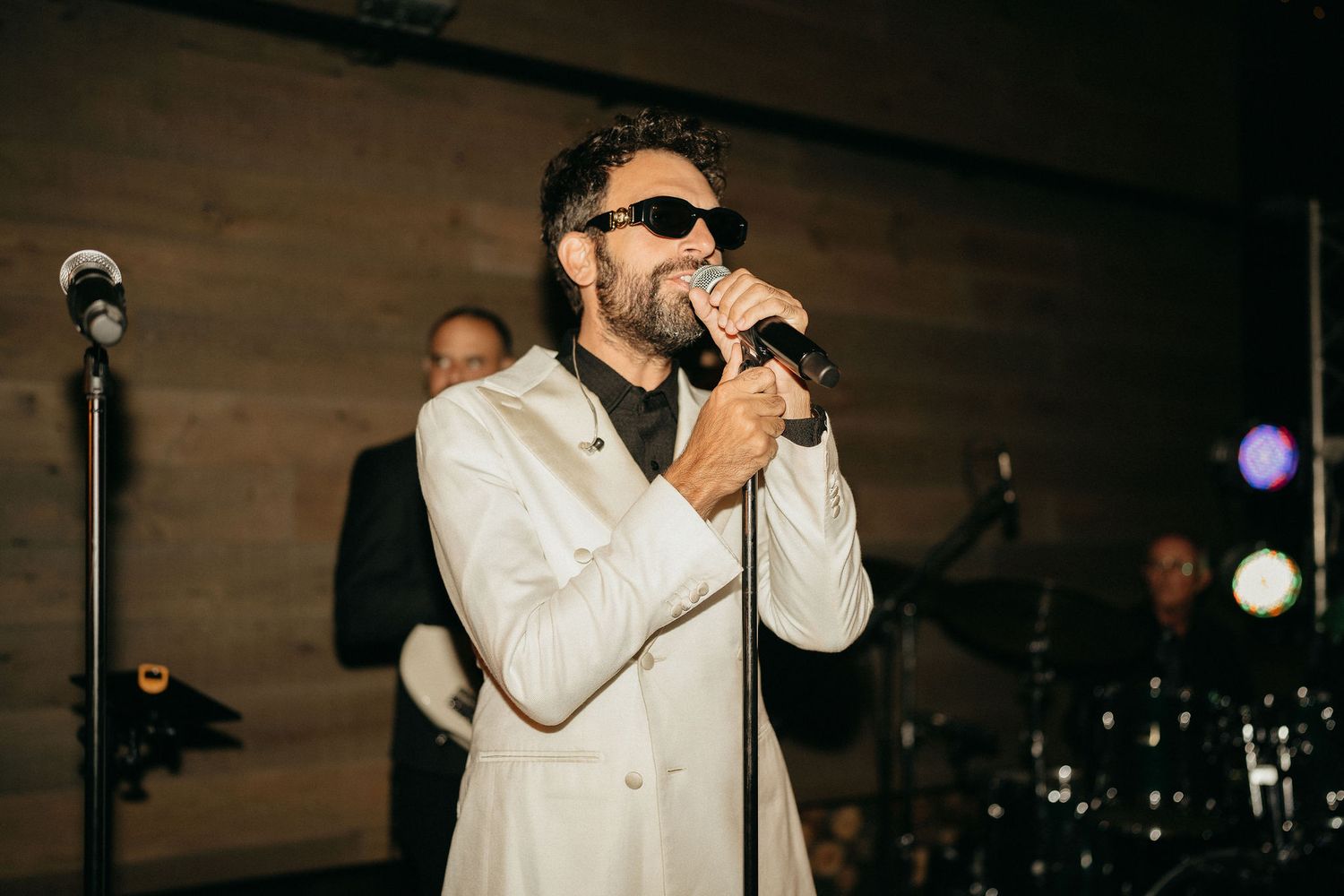 A performer in a white suit and sunglasses sings into a microphone on stage during a concert performance.