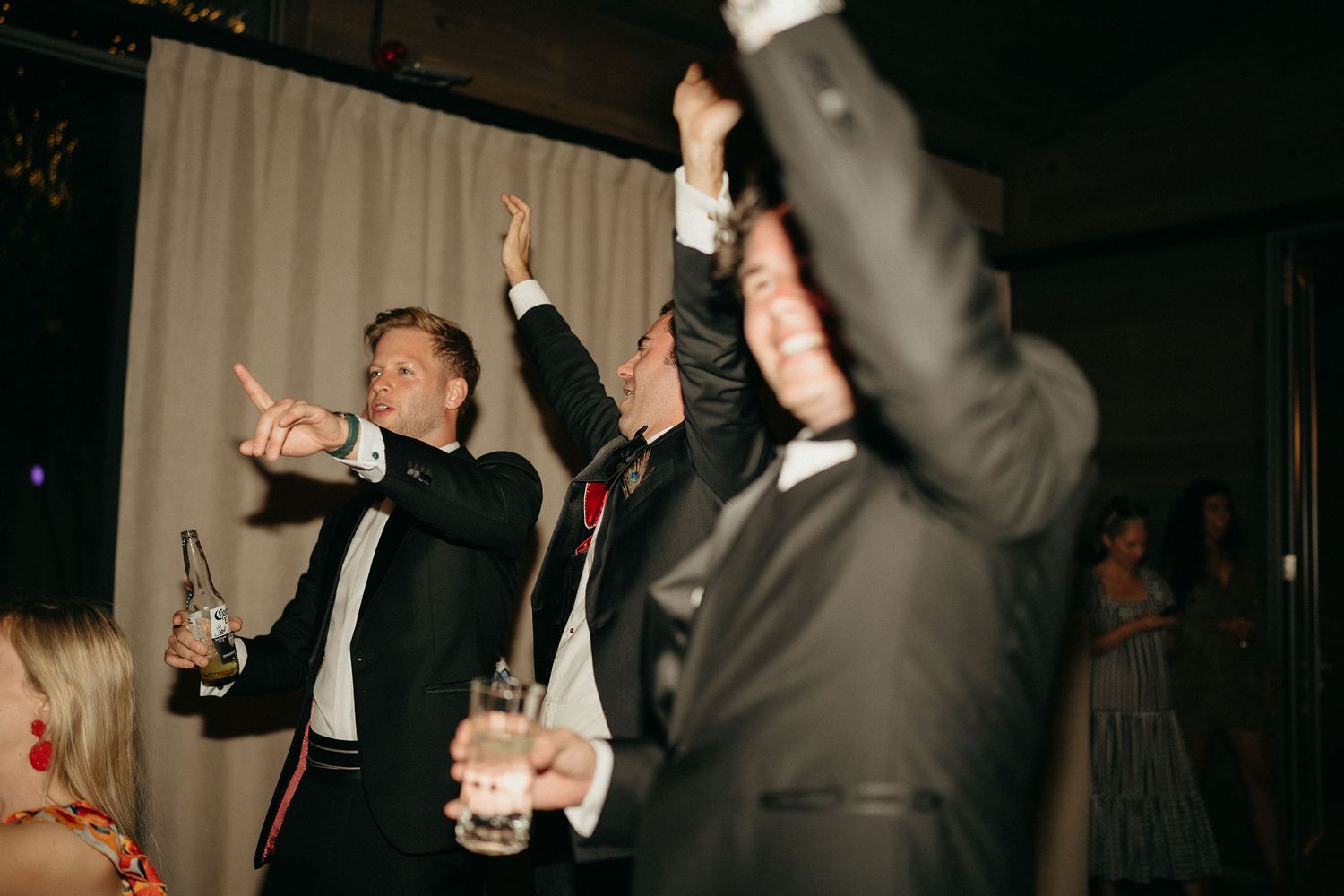 People in formal attire dancing and celebrating at a dark indoor party venue with raised hands.