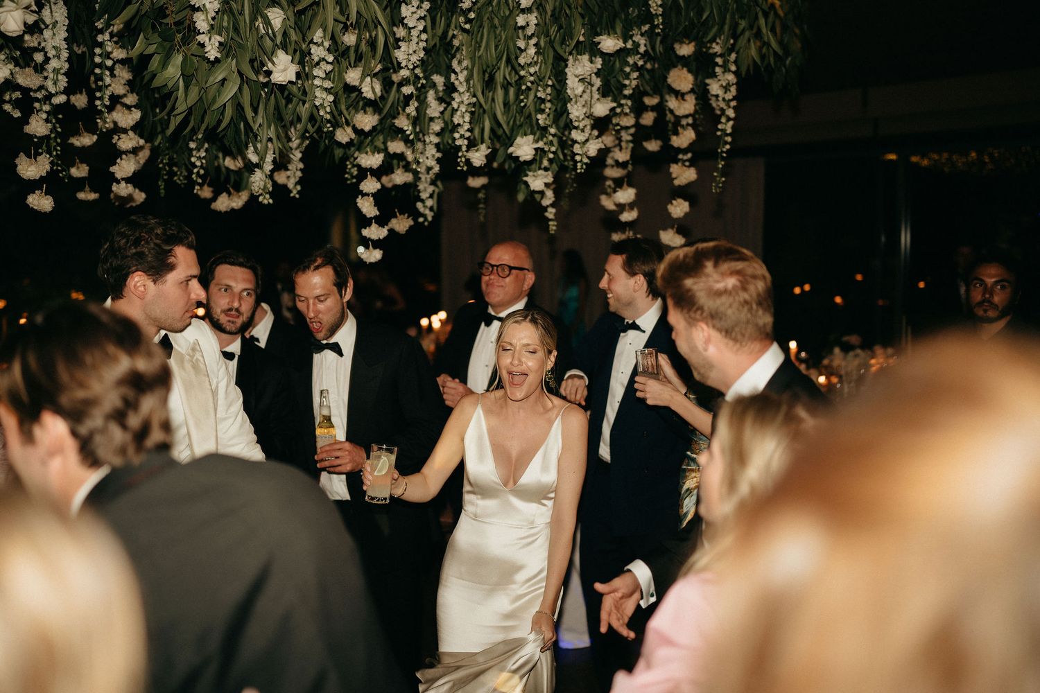 A group of wedding guests dance together on a dimly lit dance floor beneath decorative hanging lights.