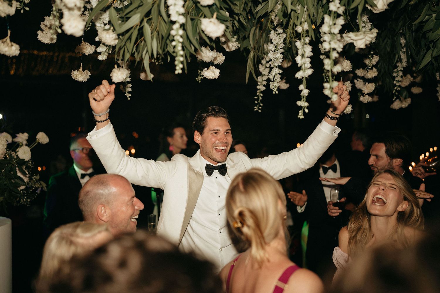 A joyful crowd dances and celebrates at an elegant wedding reception under hanging floral decorations.