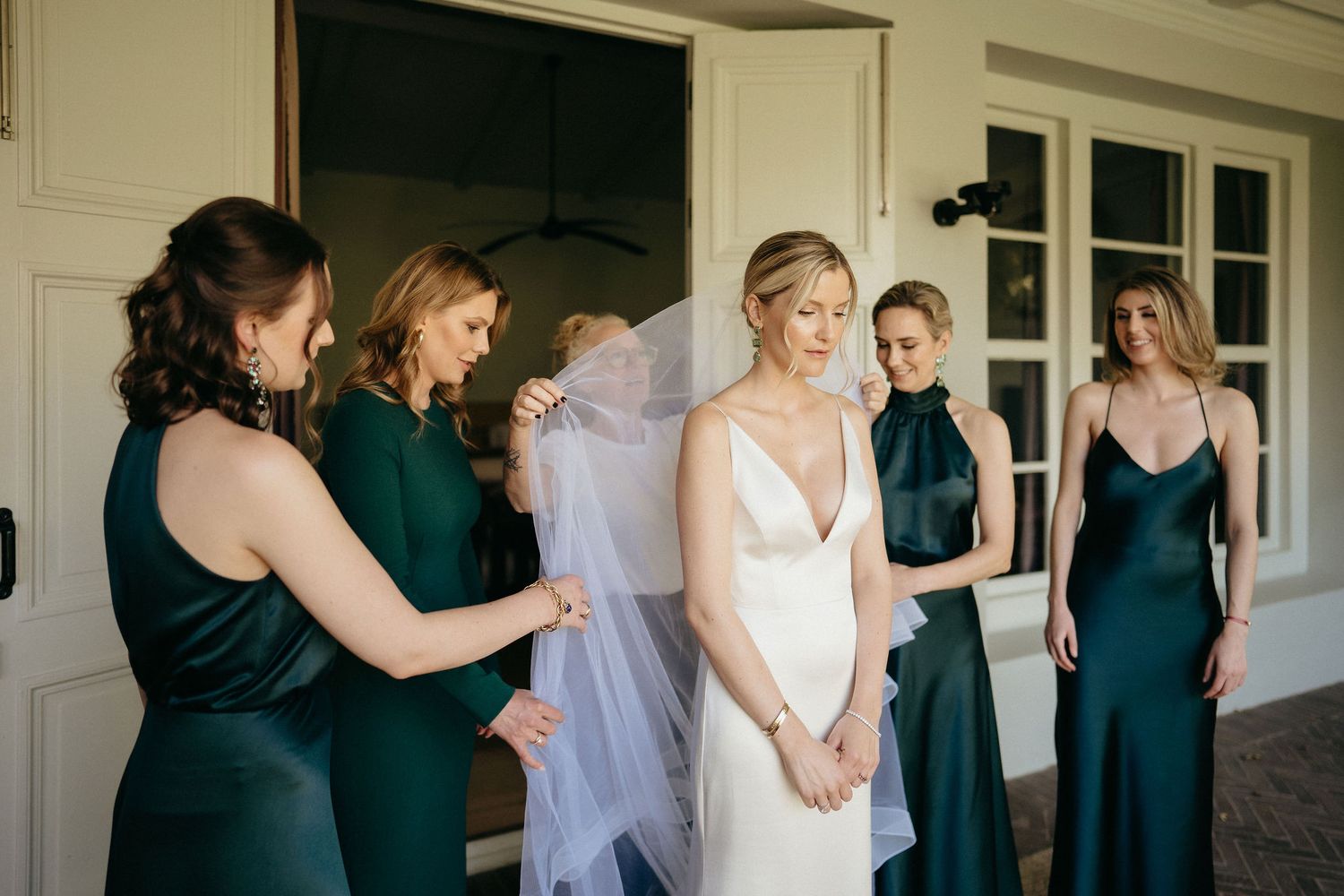 Wedding party helps bride with final preparations in a white room with natural lighting.
