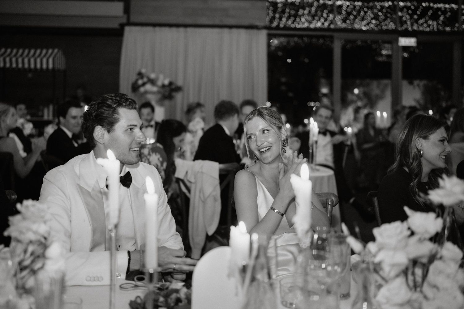 Black and white photo of guests enjoying an elegant wedding reception dinner under twinkling lights.