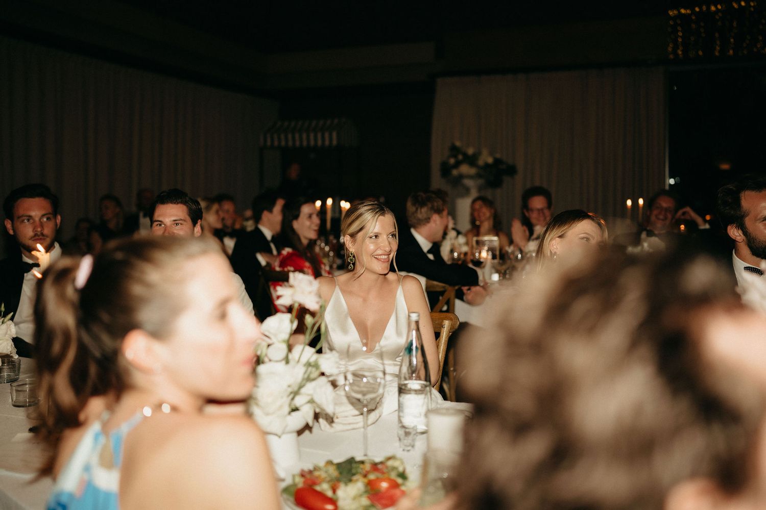 Wedding reception guests seated at round tables in a large banquet hall with white tablecloths and decorations.