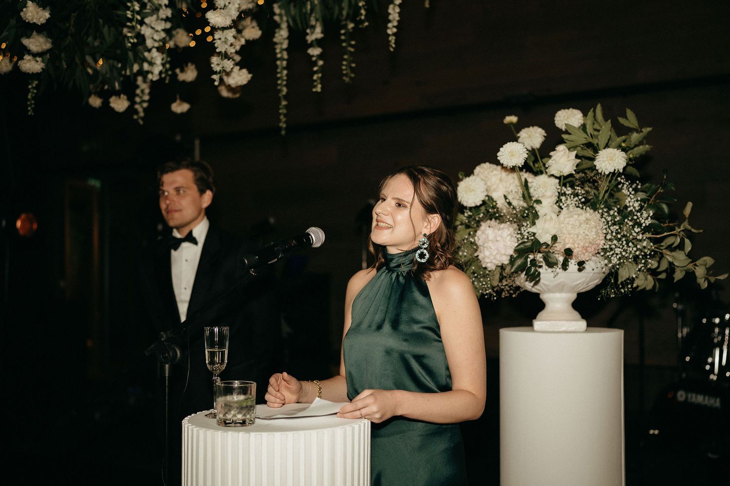 Two people in formal attire stand at a reception table next to an elegant white floral arrangement.