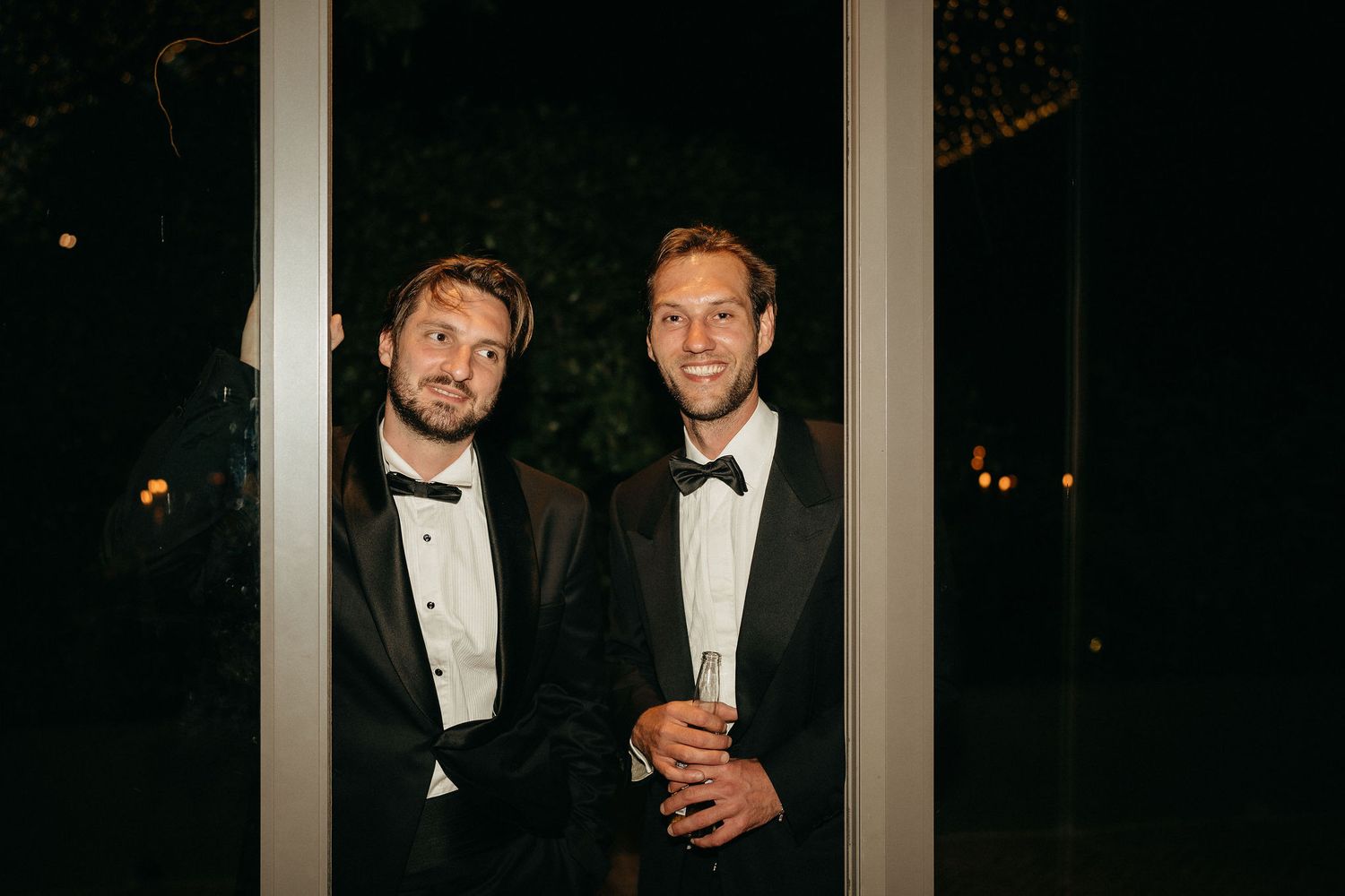 Two men in black tuxedos and bow ties stand together at a formal event in the evening.