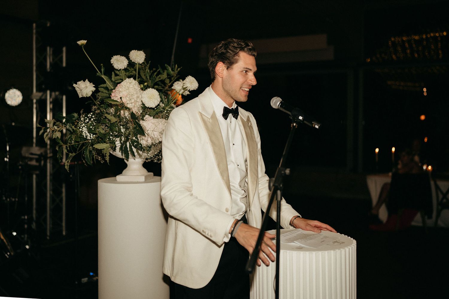 A couple dances together during their wedding reception in a dimly lit venue decorated with white floral arrangements.