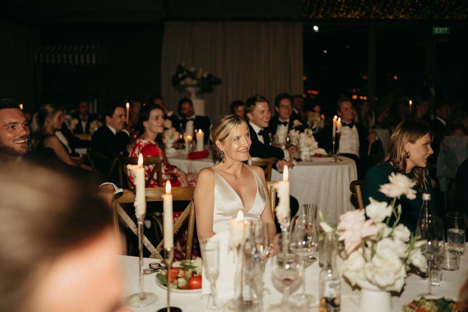 Wedding reception guests seated at elegant tables with white floral centerpieces in a dimly lit ballroom.
