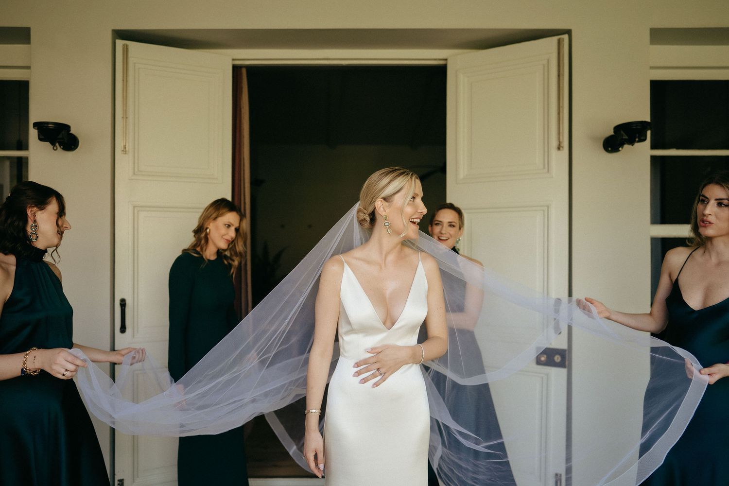 Wedding veil flows gracefully as bridesmaids assist with bridal preparations in an elegant setting.