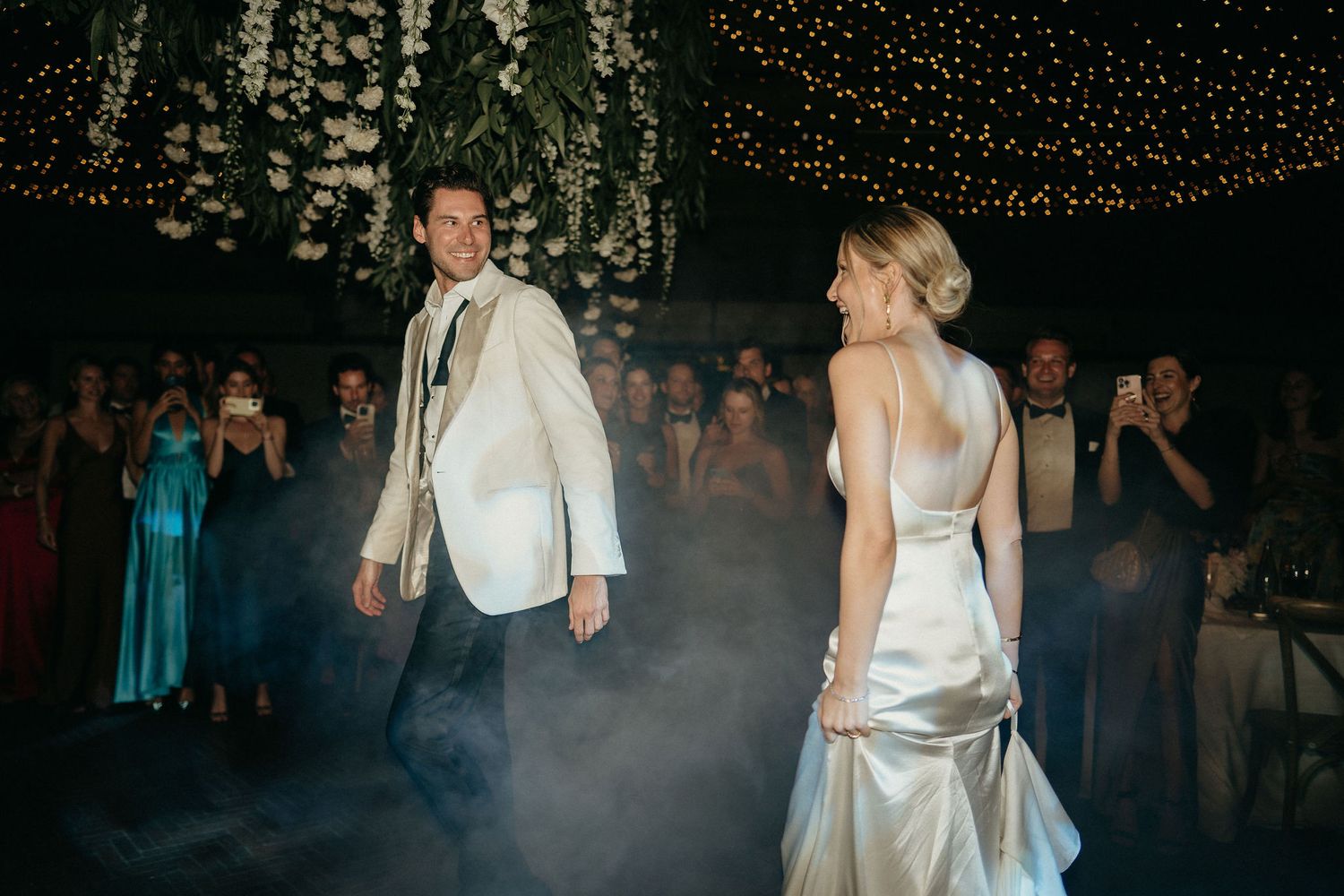 Wedding guests dance and celebrate on a dimly lit dance floor decorated with string lights overhead.