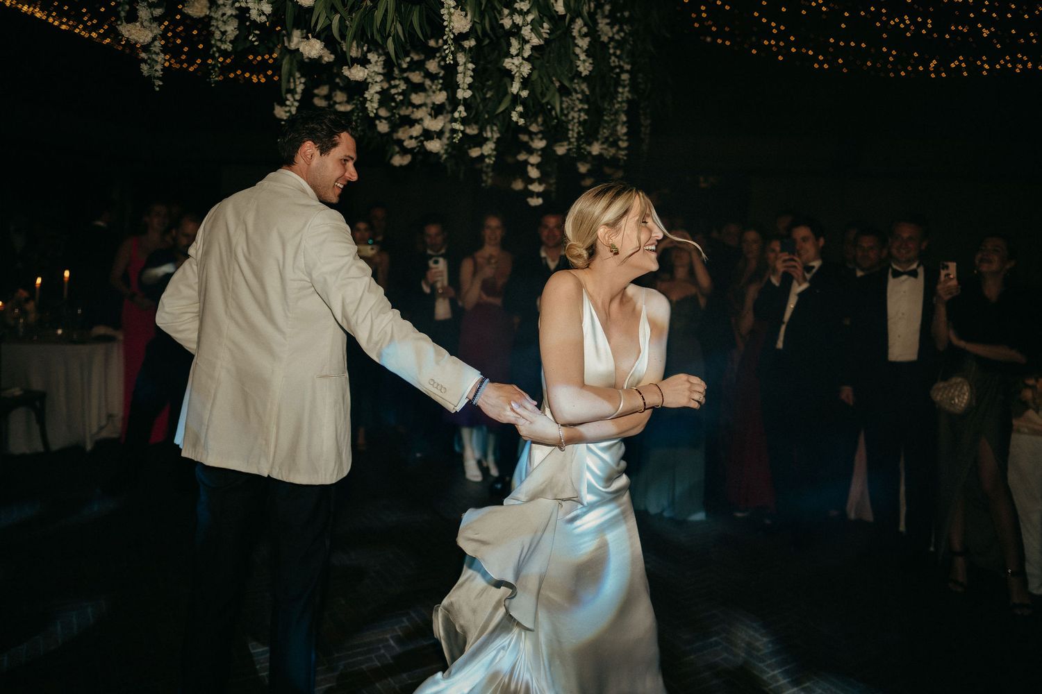 A couple shares their first dance at a wedding reception in a dimly lit venue with guests watching in the background.