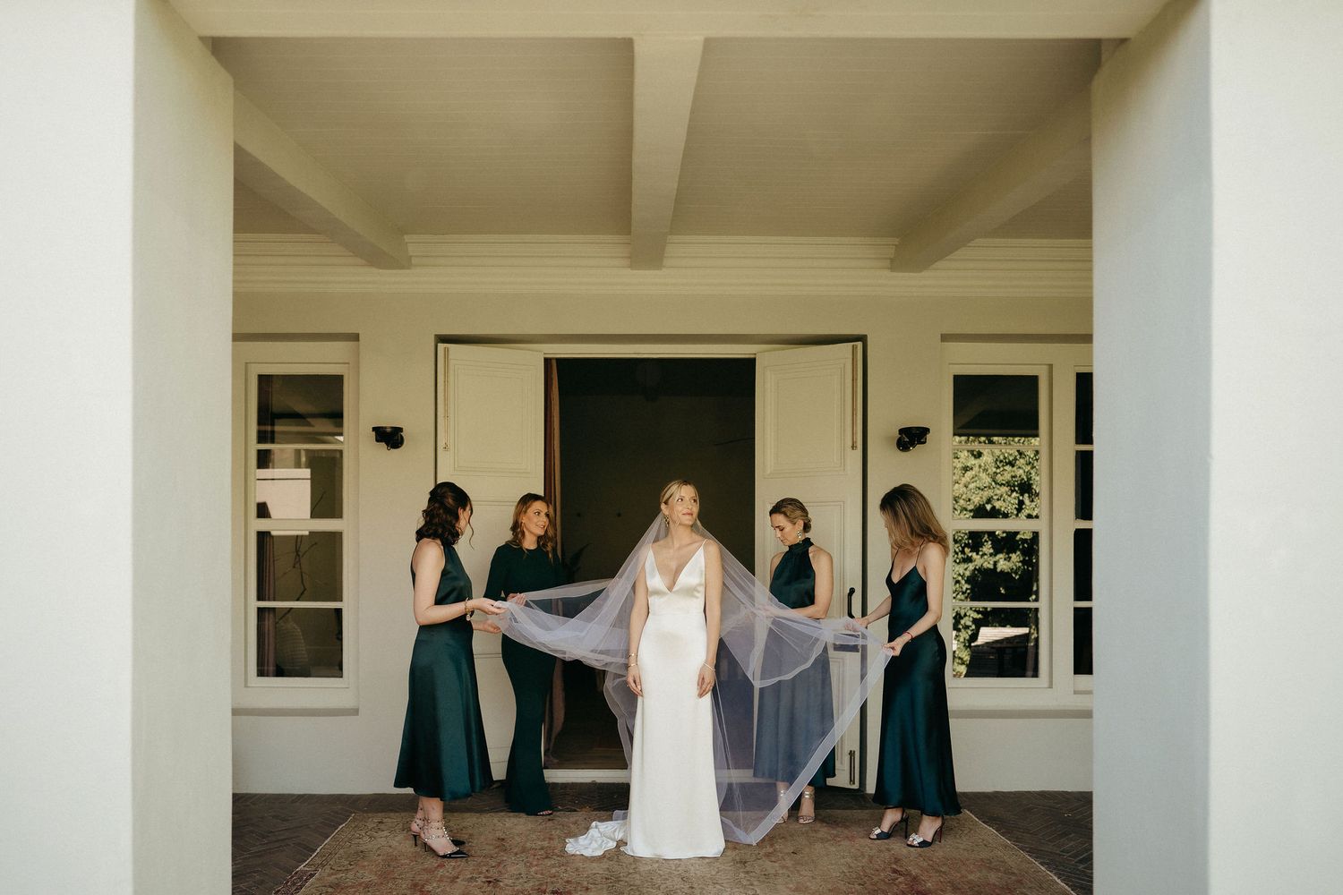 Wedding party members in teal dresses and a white gown pose in an elegant covered outdoor walkway.
