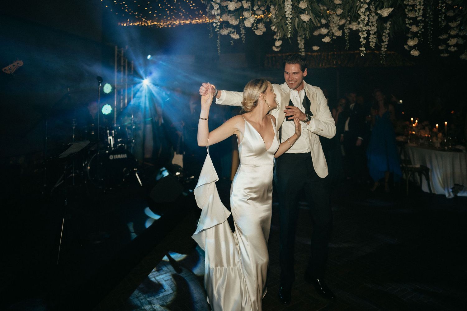 Couple shares their first dance at an evening wedding reception with blue lighting and forest backdrop.