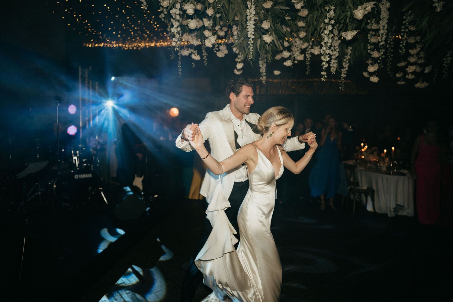 A couple dances elegantly in white formal attire at a dimly lit event with sparkling lights in the background.