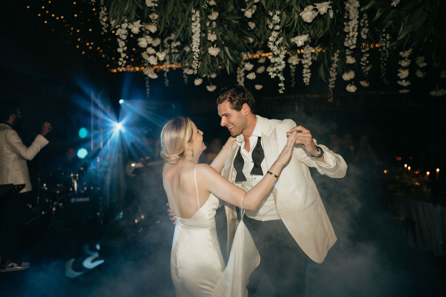 A couple dances together during their first dance at a nighttime wedding reception with twinkling lights in background.