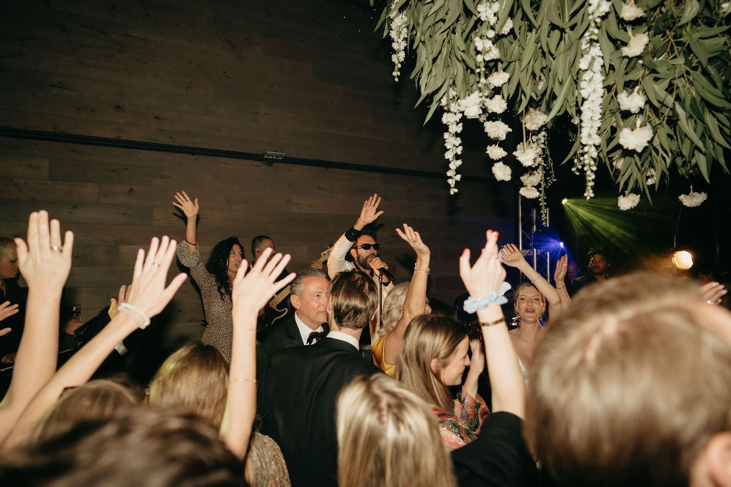 Wedding guests raise their hands in celebration on a dimly lit dance floor beneath hanging floral arrangements.