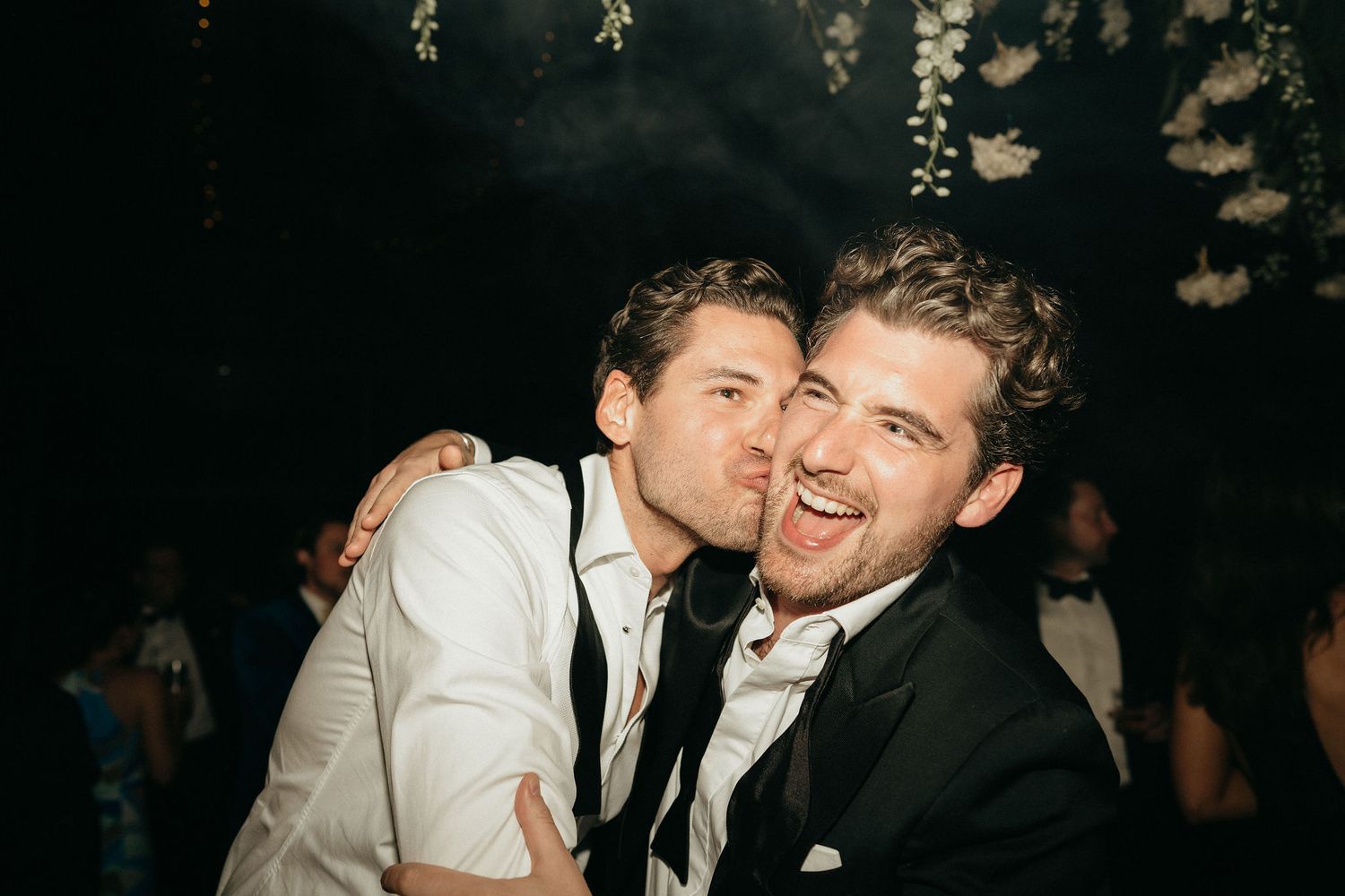 Two people in formal attire share a joyful moment at a celebration, with one leaning in close to the other.