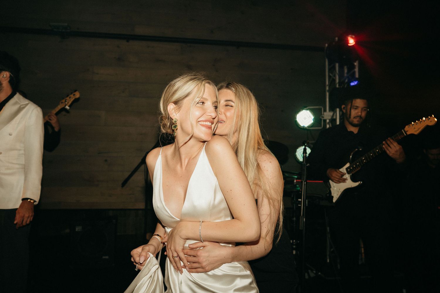 Two people in white dresses embrace on a dark dance floor while band members with guitars perform in the background.