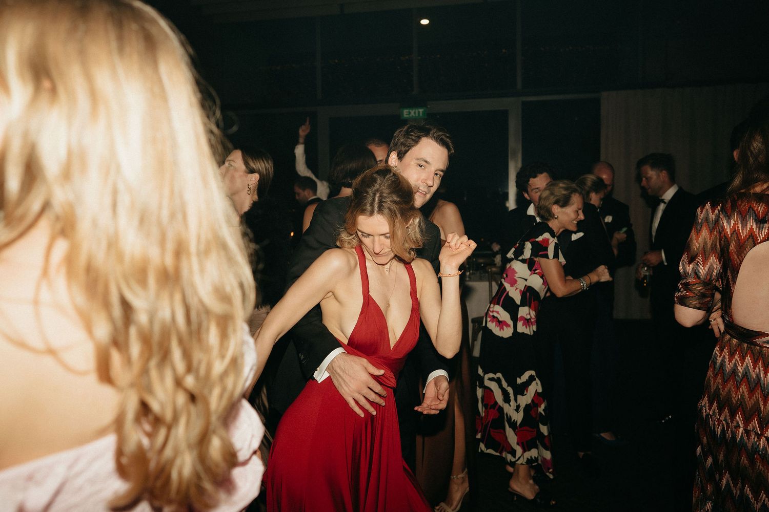 People dance and celebrate at a dimly lit formal event with red and black attire visible in the crowd.
