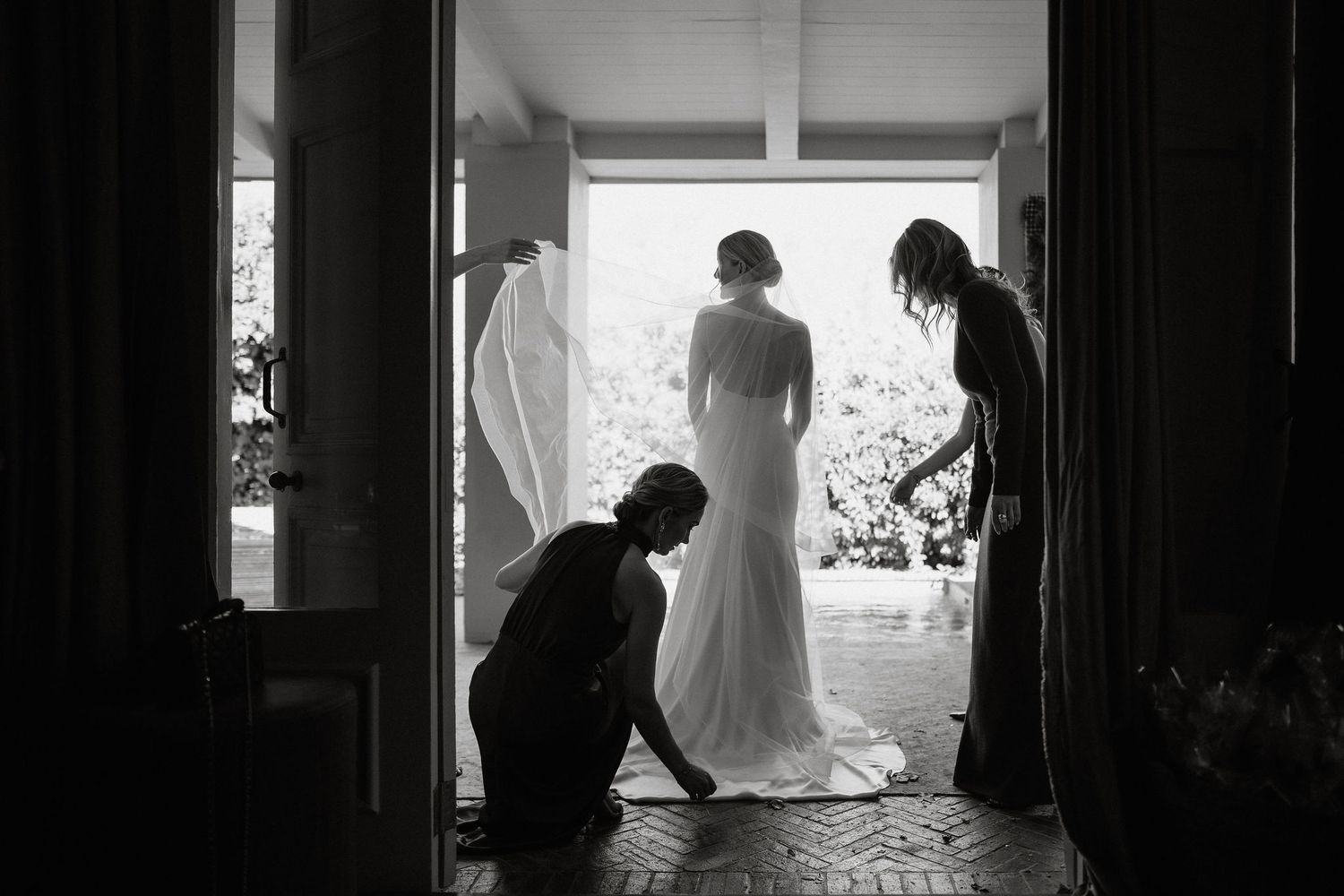 A black and white photograph shows a wedding dress being adjusted by silhouetted figures in a doorway.