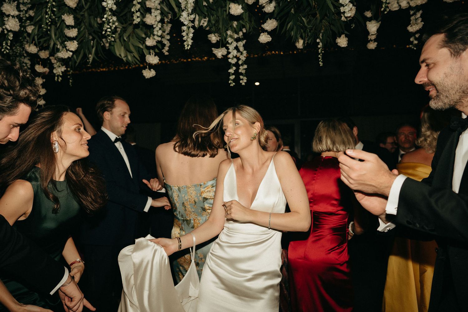 Wedding guests dancing and celebrating on the dance floor under hanging floral decorations at an evening reception.