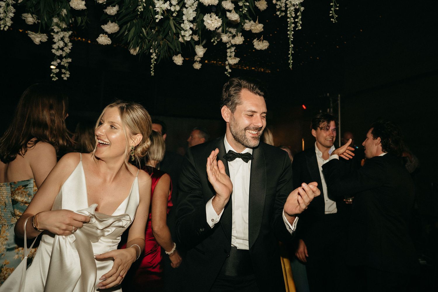 People dancing and celebrating at a dimly lit party or reception with decorative lights hanging from above.