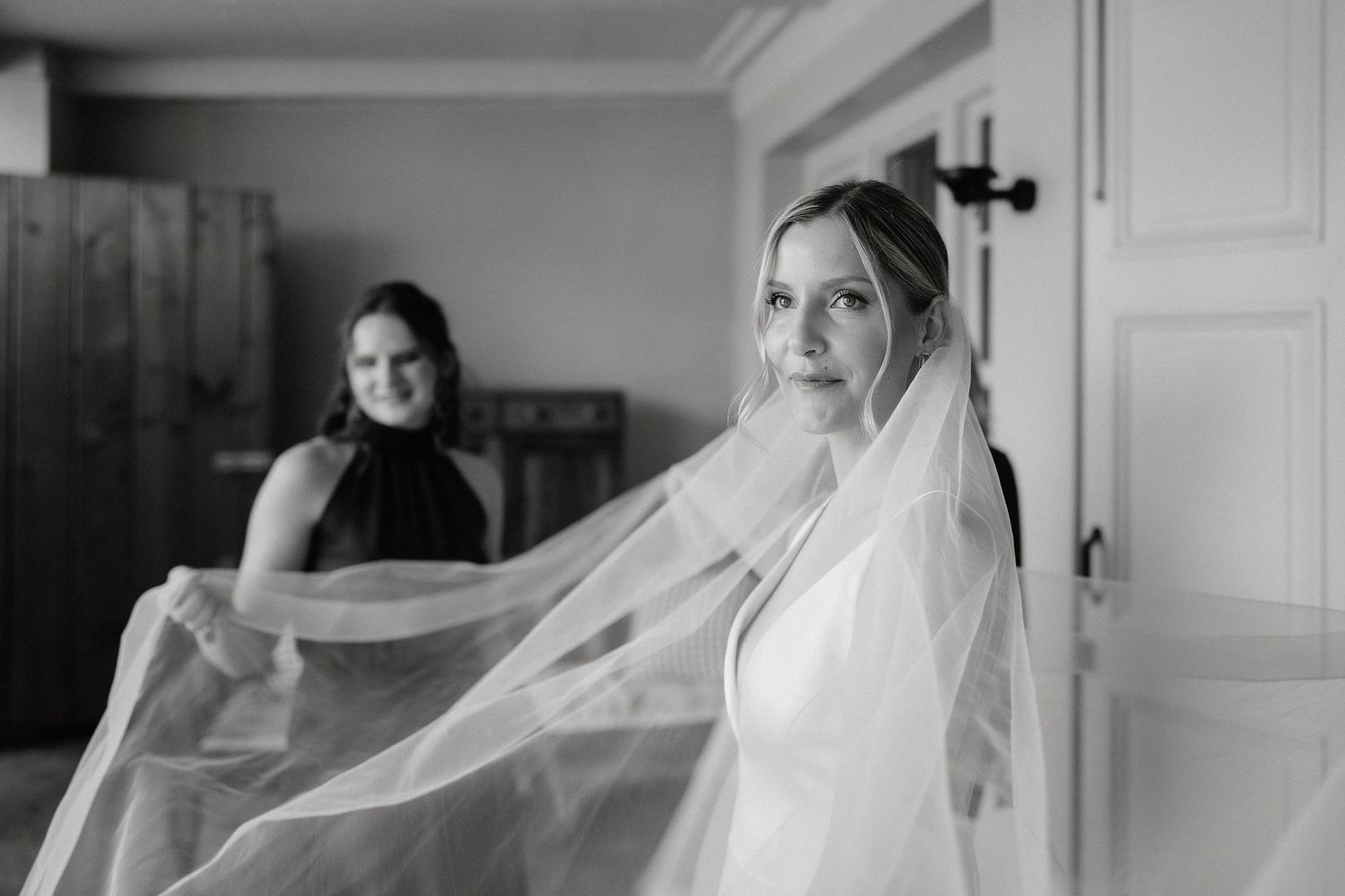 Black and white photo of a bride wearing a flowing veil while another person in dark dress stands in background.