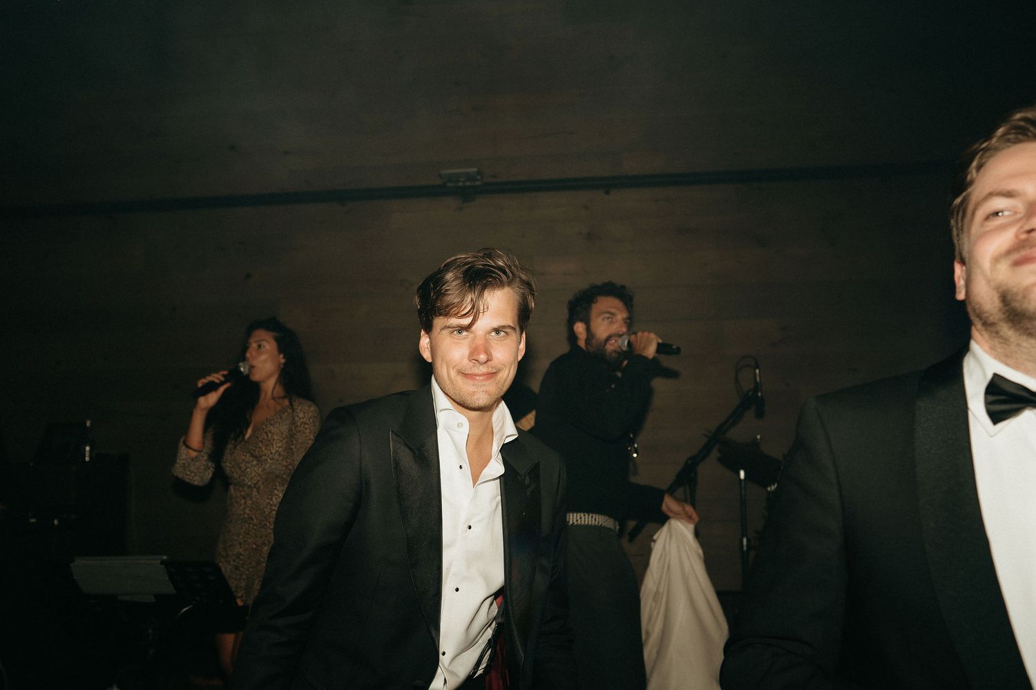 People in formal attire gather in a dimly lit room during what appears to be an evening event or celebration.