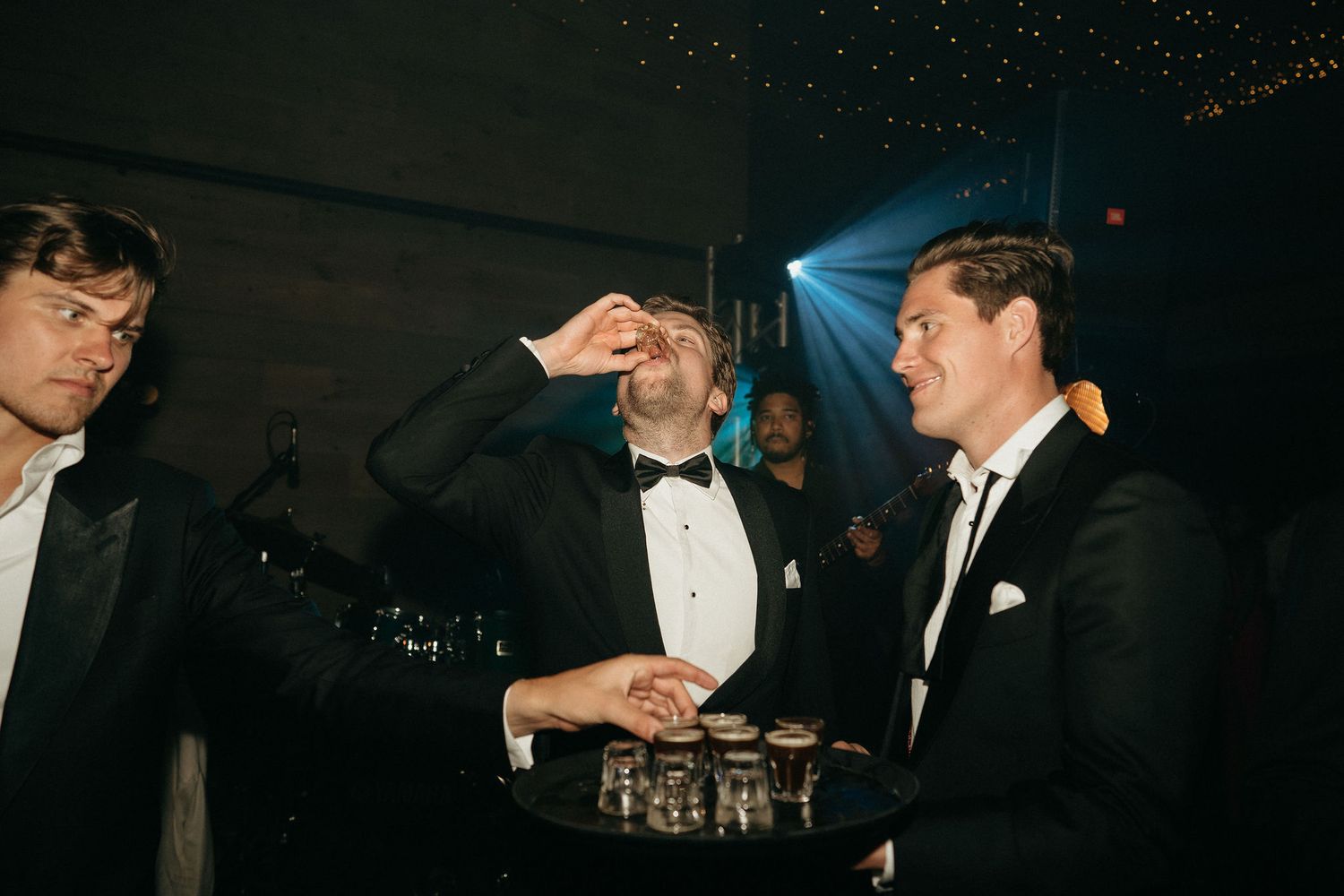 Three people in formal attire with bow ties interact at an evening event while holding drinks.